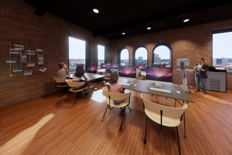 This photo is a rendering of the digital art studio located on the second floor. There are four workstations, each with two computers for students to use, and a computer station set up for the instructor at the front of the room, in front of large printer. There is an instructor at the front of the room and few students working on the computers. There are windows on each of the two surrounding walls, and some printed out photos hung up on the side wall.