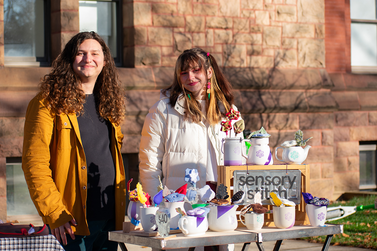 Two design students stand behind their market display table, which features an assortment of colorful, handmade, sensory fidget objects.