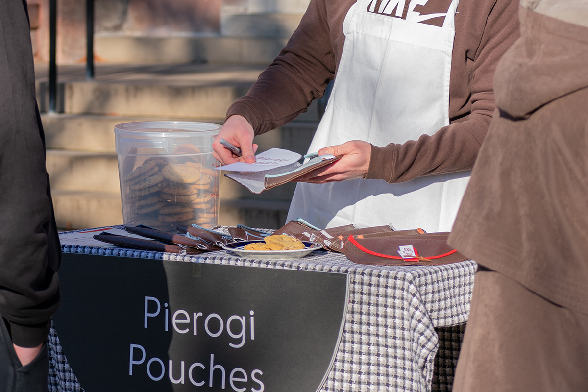 A person’s hands are shown unfolding a Pierogi Pouch product, which looks like a half-circle made from dark and light-colored fabric-like material, to reveal a care and use card inside; the person stands behind a market display table arranged with more products and a bucket filled with cookies.