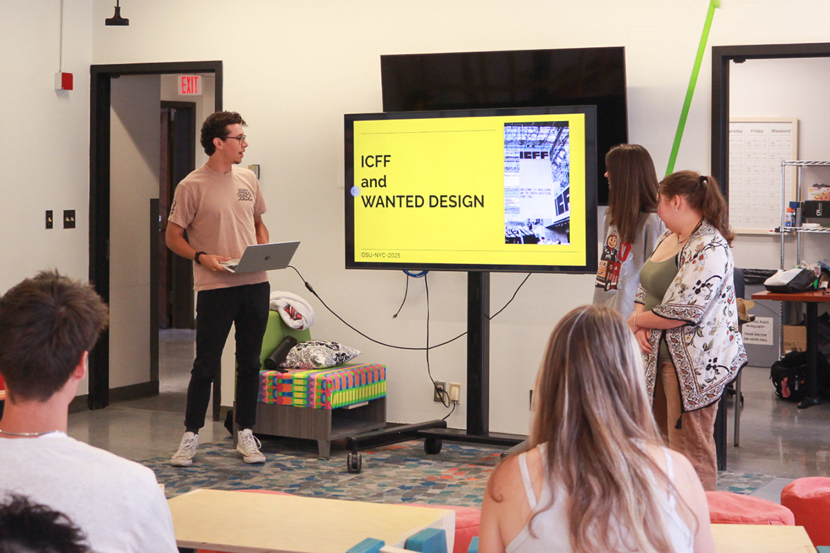 Photo of three students presenting next to a TV in front of a classroom full of students.