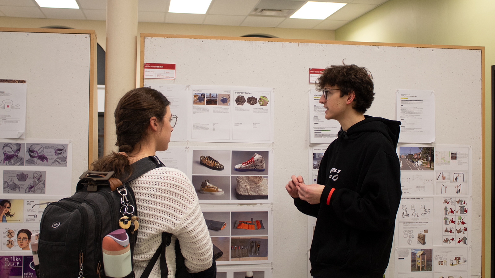 Two people stand in front of a display of industrial design project boards. One person explains the work on the board.