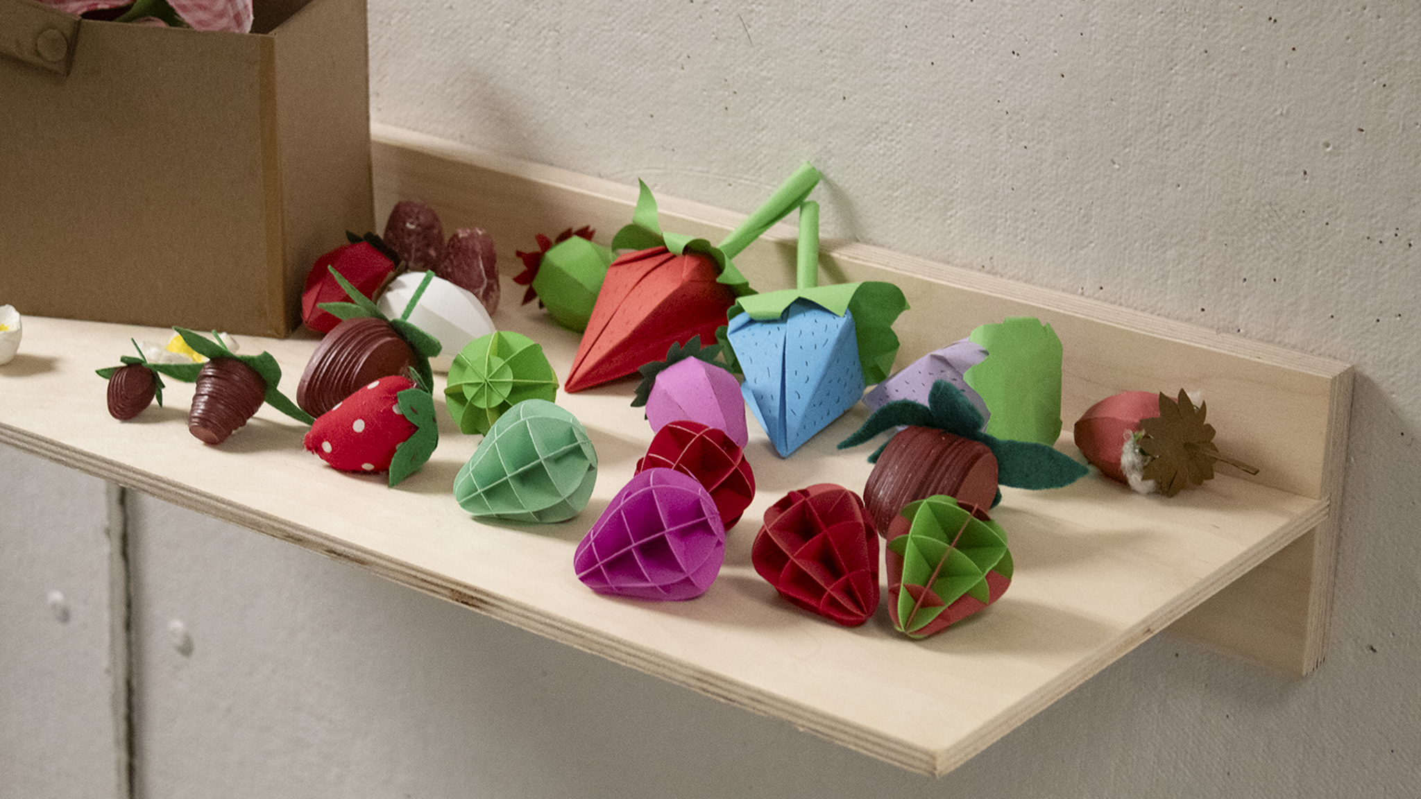 Wooden wall shelf displaying a variety of colorful handmade strawberry crafts in diﬀerent materials, textures, and colors, with more pieces in a box on the left