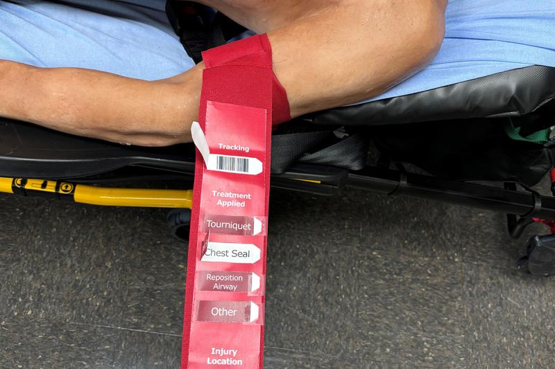 A red tag hanging off of an arm labeled with “tracking, “treament applied,” “tourniquet,” “chest seal,” “reposition airway,” “other,” “injury location,” and a clip art styled person. The “tracking” pull tab reveals white underneath. The “chest seal” tab reveals white underneath with black lettering