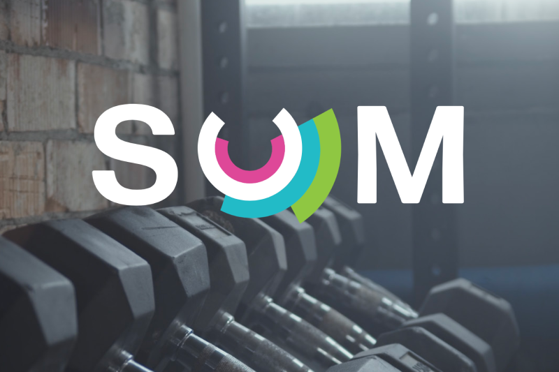 The SUM logo in front of a dumbbell rack