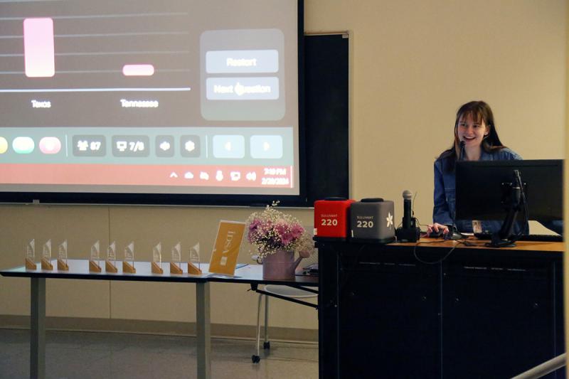 a woman speaking at a lectern next to a projector screen with trivia on it