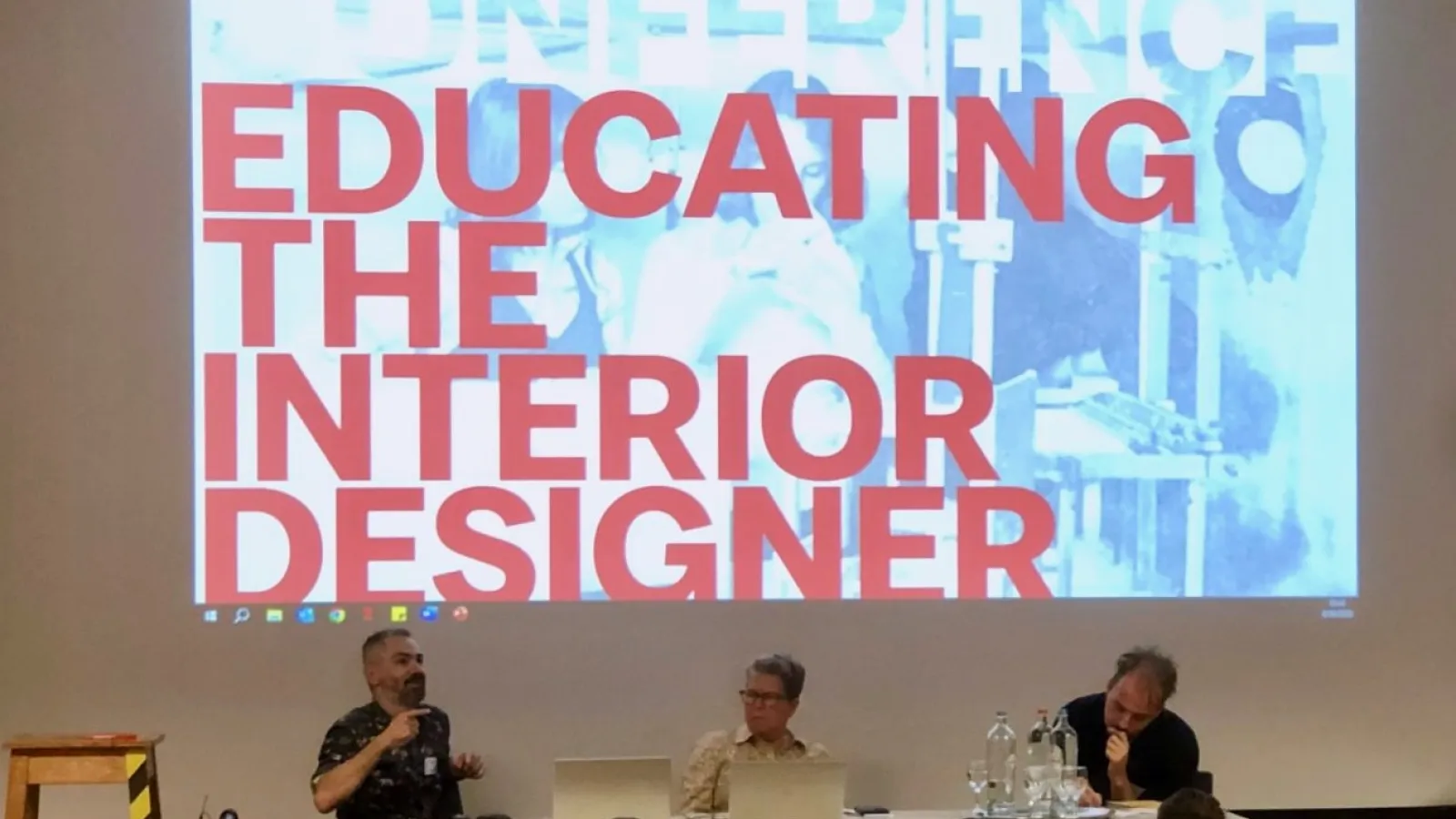 Panelist seated at a table in front of a projection that states "Educating the interior designer."
