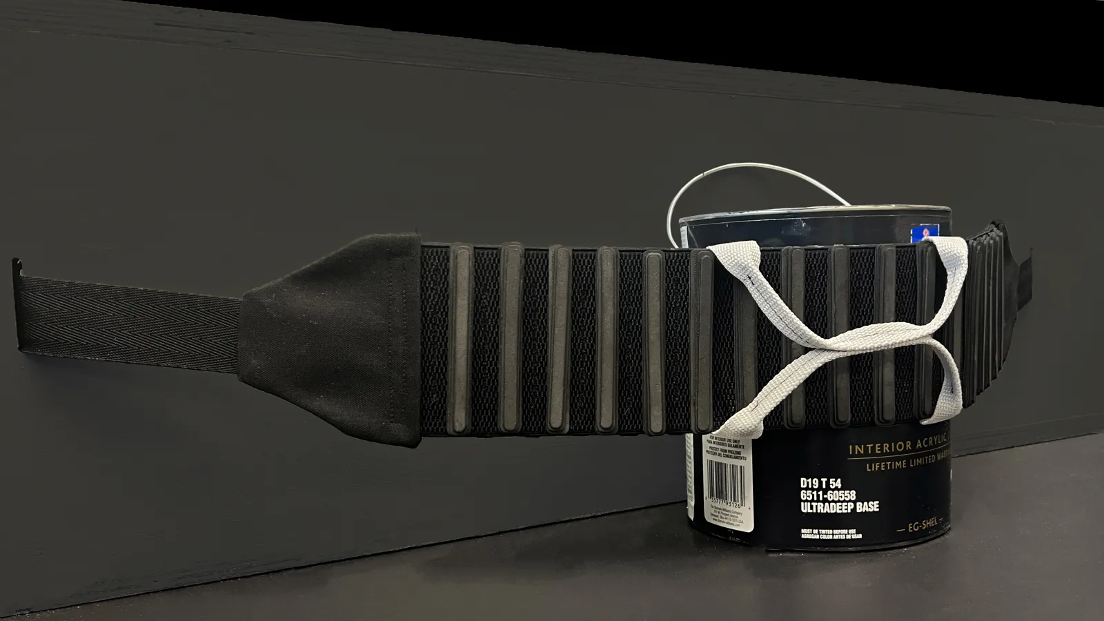 A close-up view of the FrunkFlex cargo-securing system stretched around a paint can. The black elastic strap features ribbed knit padding and a light gray activation handle, demonstrating how it conforms to cargo. The background is a black surface, emphasizing the product’s structure and functionality.