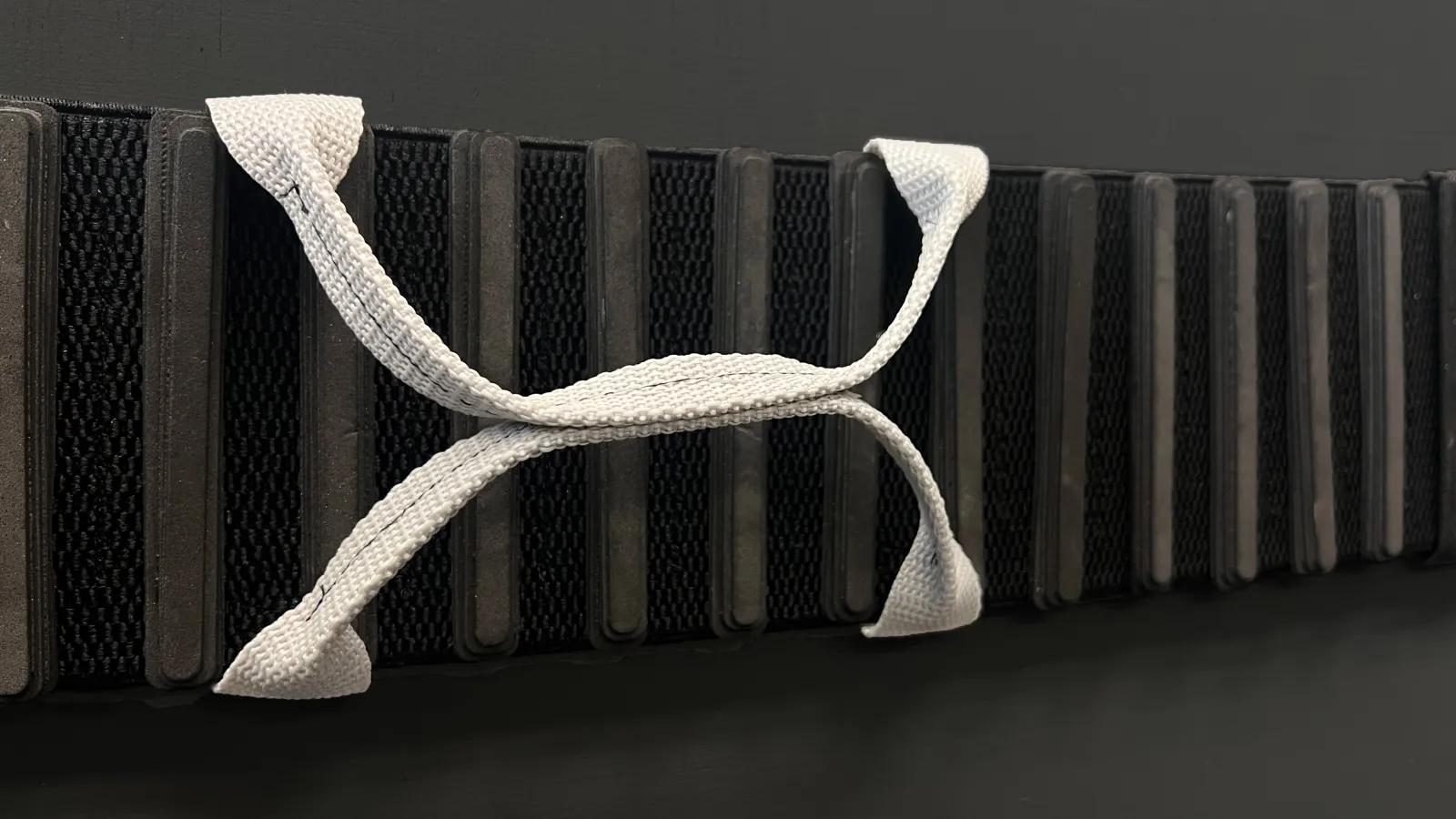 A detailed view of the FrunkFlex cargo-securing system. The black elastic strap features ribbed knit padding, with a light gray woven handle forming an "X" shape. The textured elements highlight its flexibility and grip, set against a dark background.