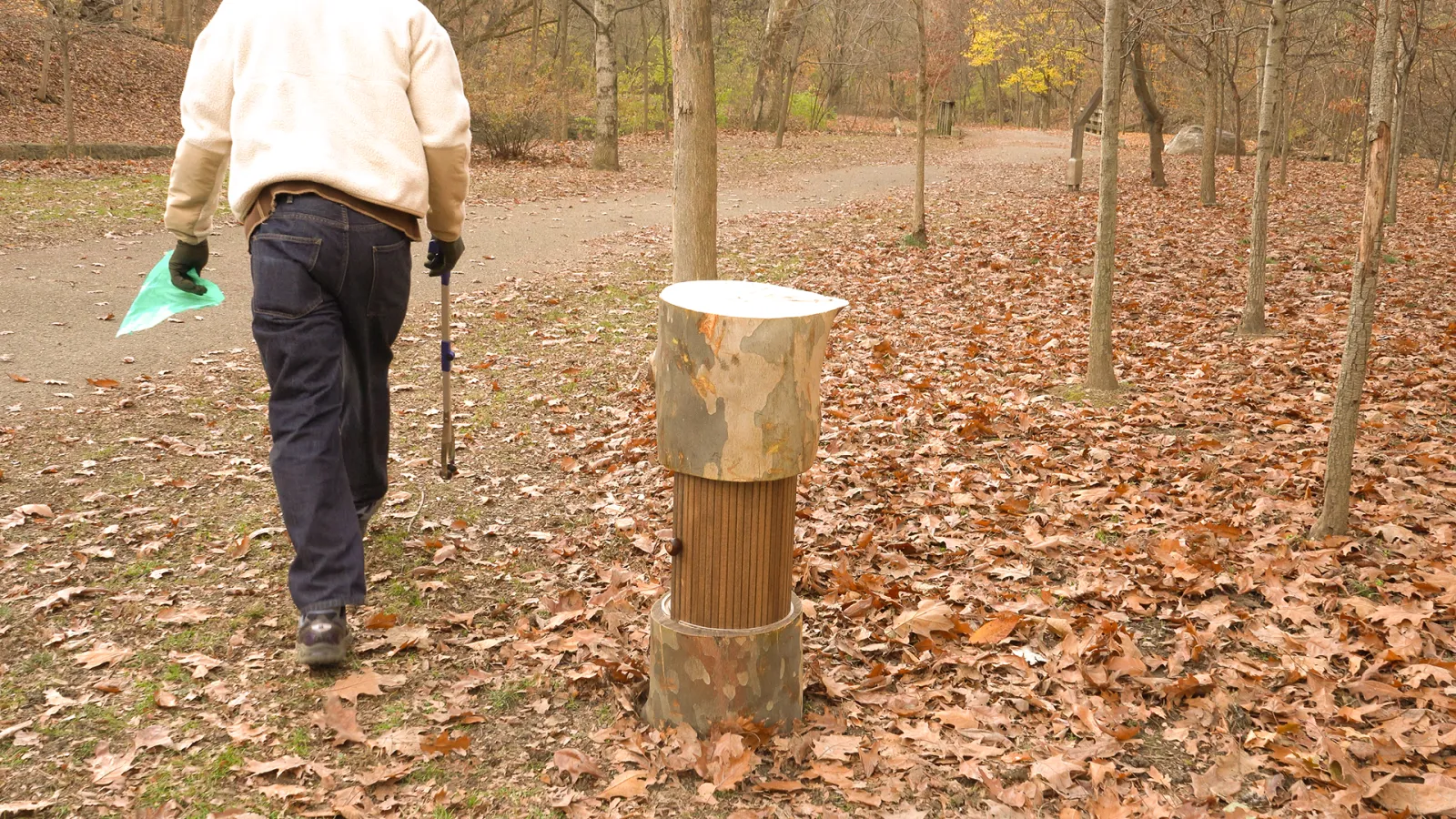 A person walks away from the tree-stump-inspired structure (Heritage Ring), headed toward a paved path with a trash grabber and trash bags.