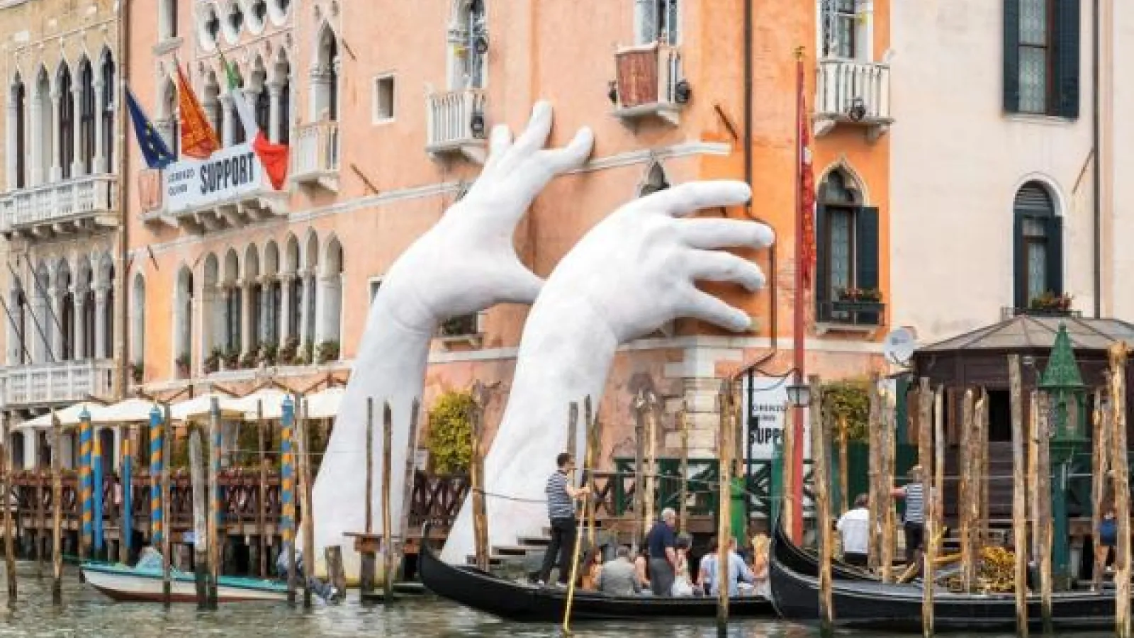 Venice art installation of giant hands coming out of water and touching a building.