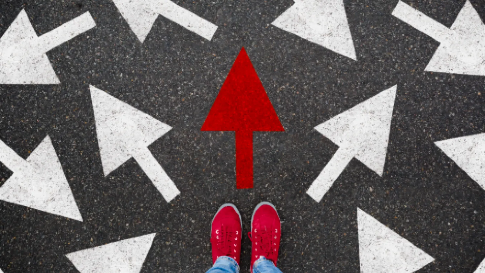 Top down view of person in jeans with scarlet shoes, standing on gray concrete with lots of white arrows pointing every which way and one scarlet arrow pointing the way forward.