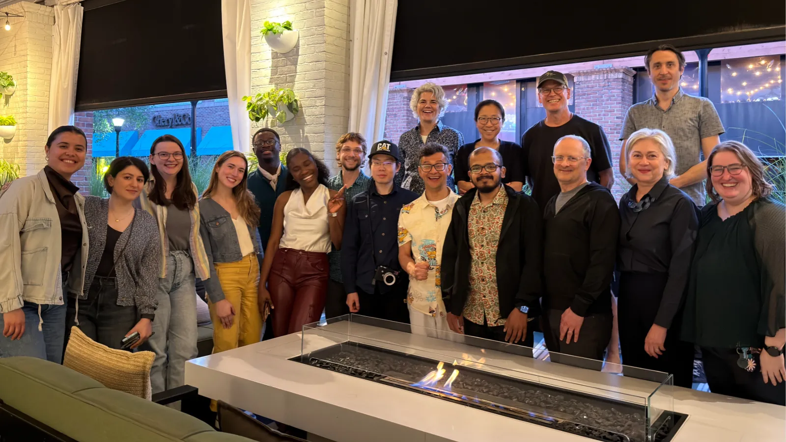 A group of students and faculty stand smiling around a fire pit