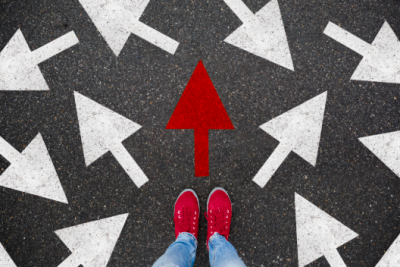 Top down view of person in jeans with scarlet shoes, standing on gray concrete with lots of white arrows pointing every which way and one scarlet arrow pointing the way forward.