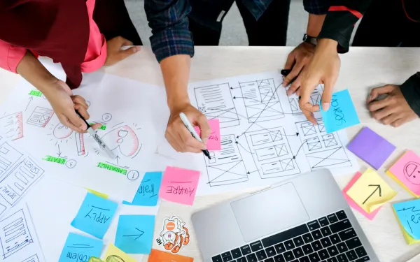 overhead view of three people collaborating at a desk with drawings, post-it notes, and a laptop