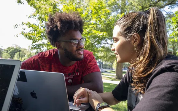 Two students outside on campus talking with a notebook computer in front of them