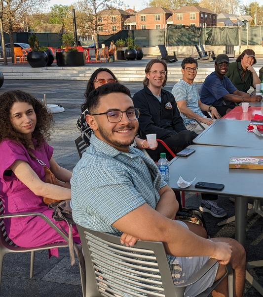 Seventeen people sitting around a long table, looking at the camera, outdoors on the OSU campus.