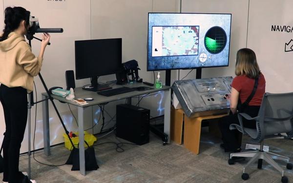 Two people in a room, one is seated working at a gray 3D interface and one is standing and looking through a VR headset.