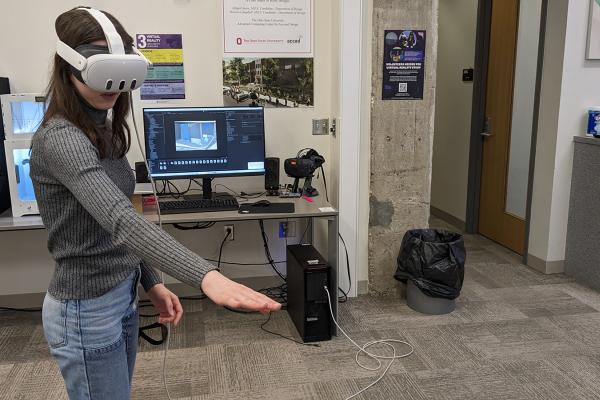 A person wearing a VR headset with their right arm extended and a screen in the background showing what the person is seeing.