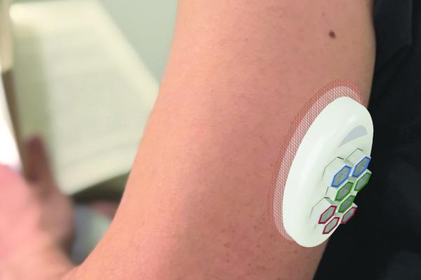 A wearable B.Well device attached to a person’s upper arm. The device has hexagonal, color-coded dosing cabins and is secured with a skin patch. The person is holding an open book, showing the device being used in a daily setting.