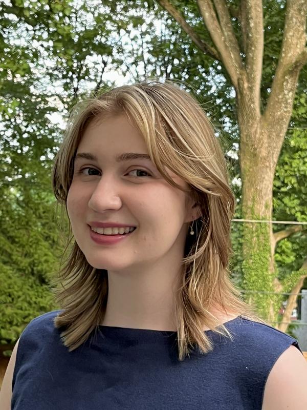 Sophia Reeder is a young woman with blonde hair. She is wearing a navy blue top and smiling at the camera. There are trees behind her.