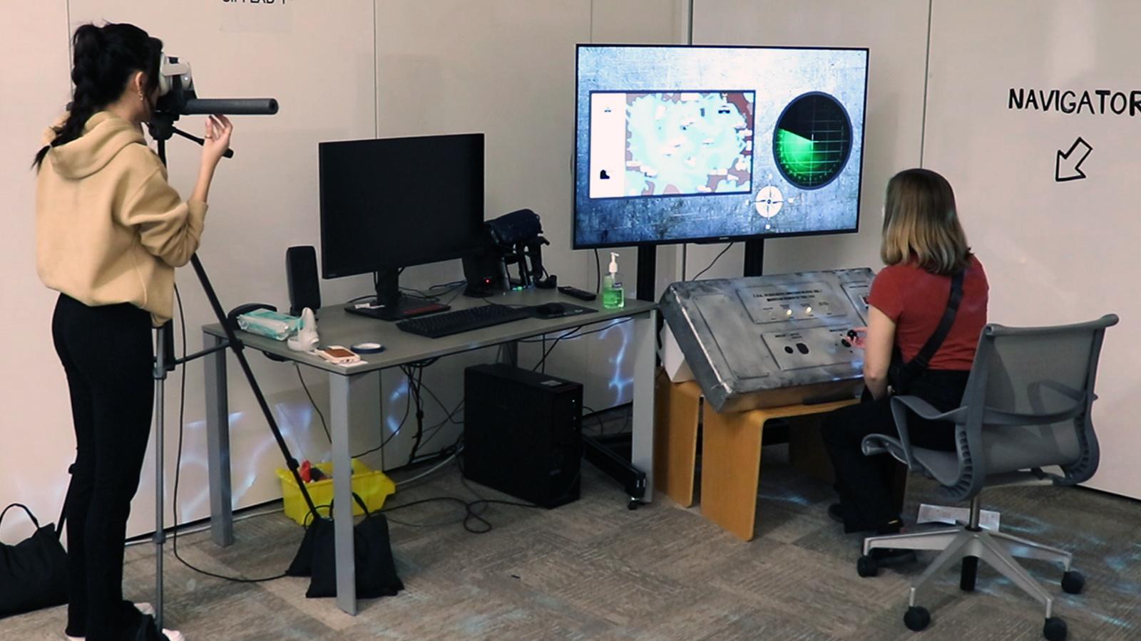 Two people in a room, one is seated working at a gray 3D interface and one is standing and looking through a VR headset.