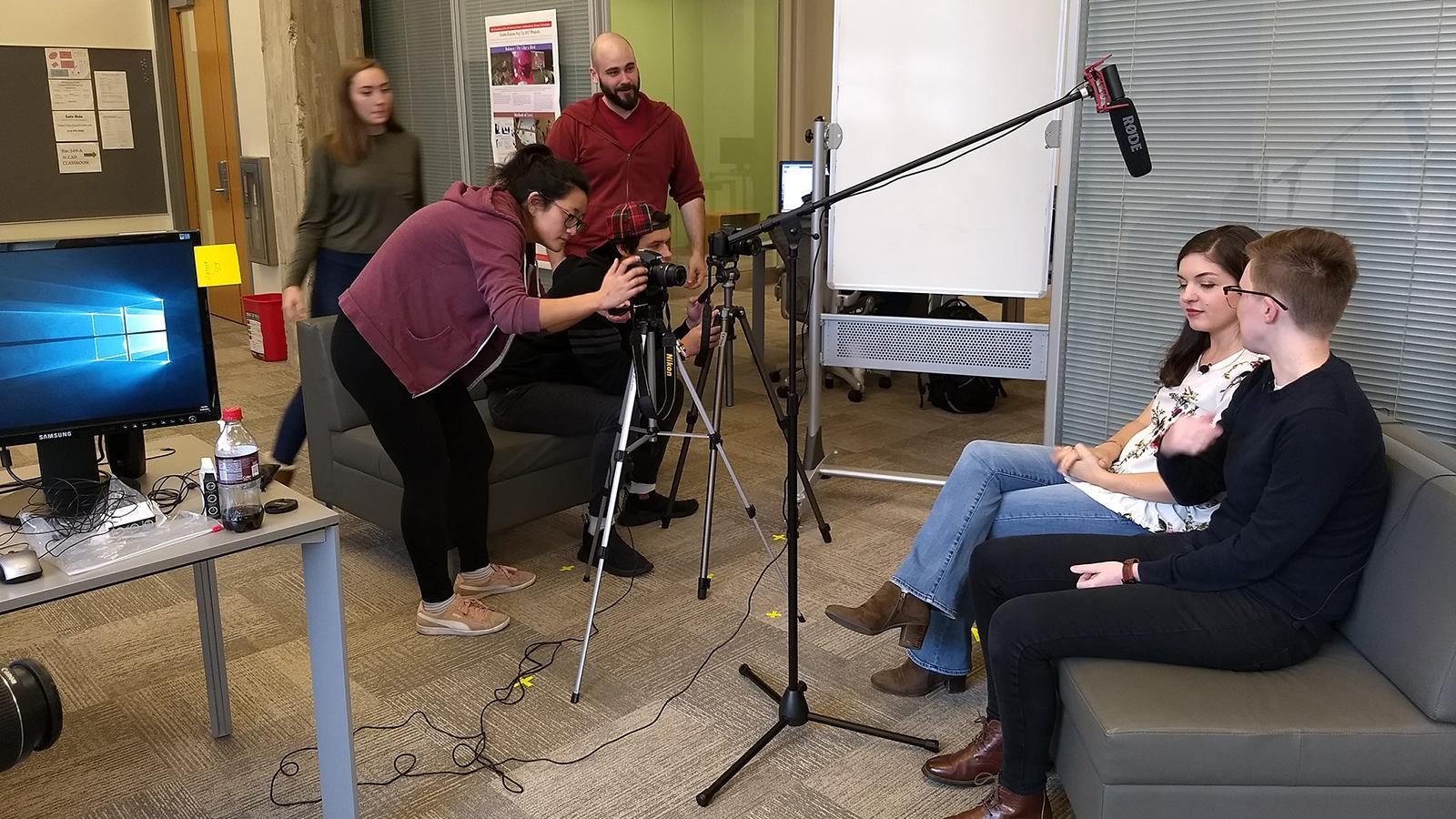 Interior room with large glass walls in which four students are aligning cameras and microphone towards two people sitting on a couch.