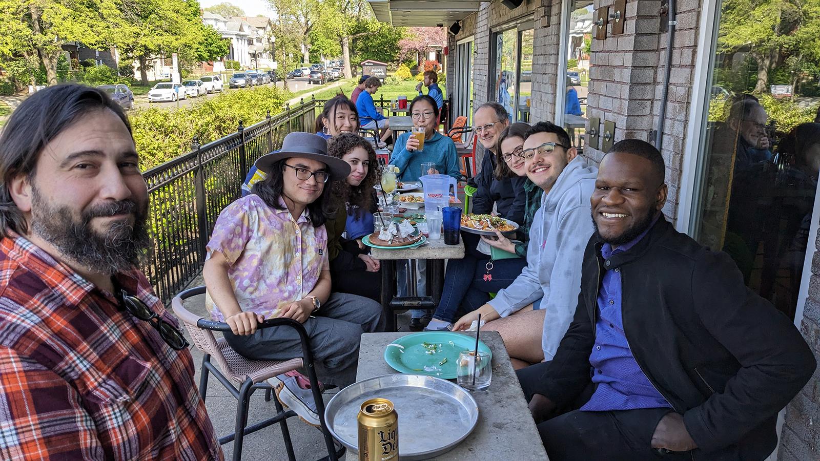Nine people looking at the camera, sitting around two tables on an outdoor porch.