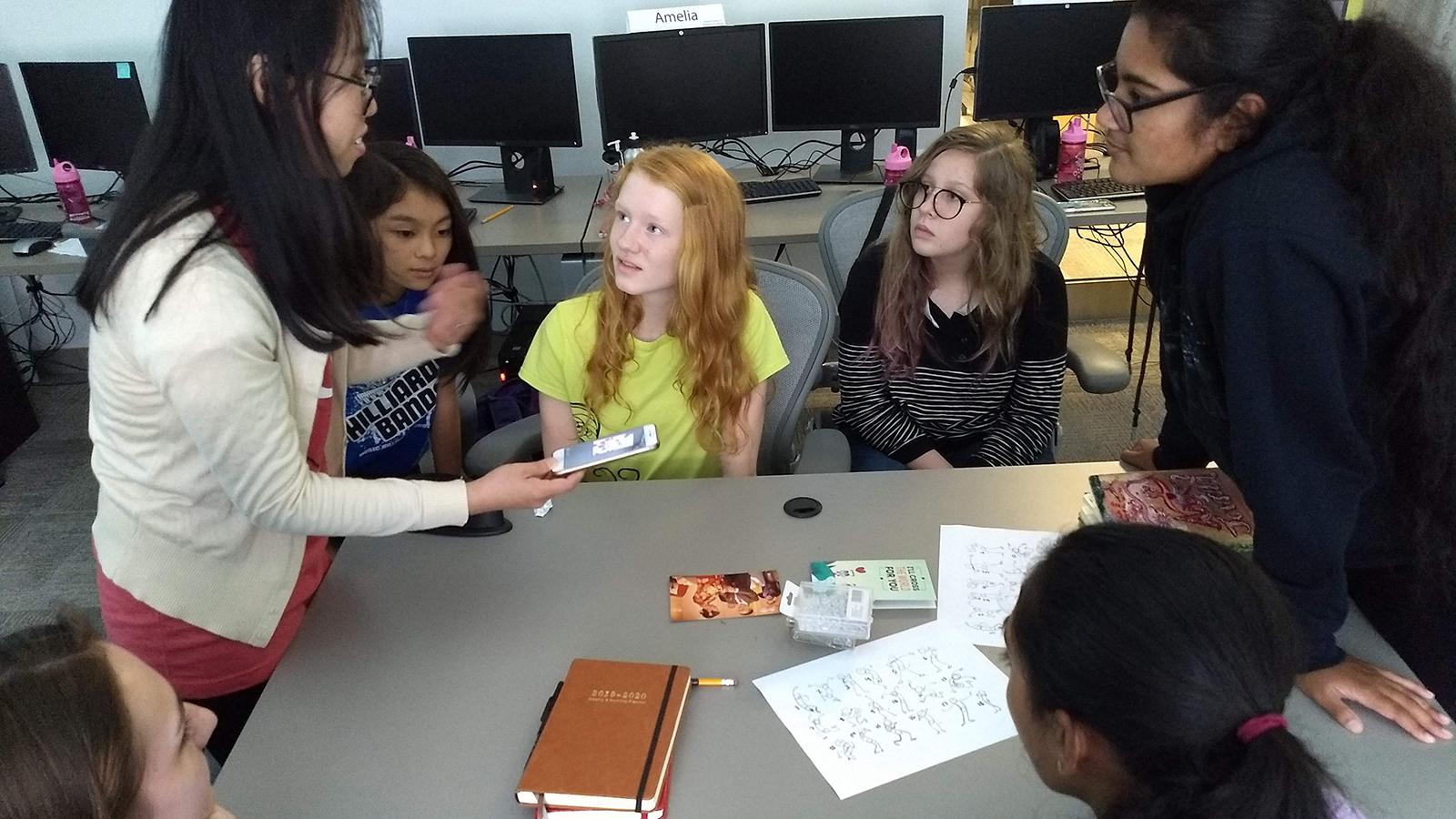 Seven people are gathered around a table, looking at the person on the left who is holding out a smart phone.