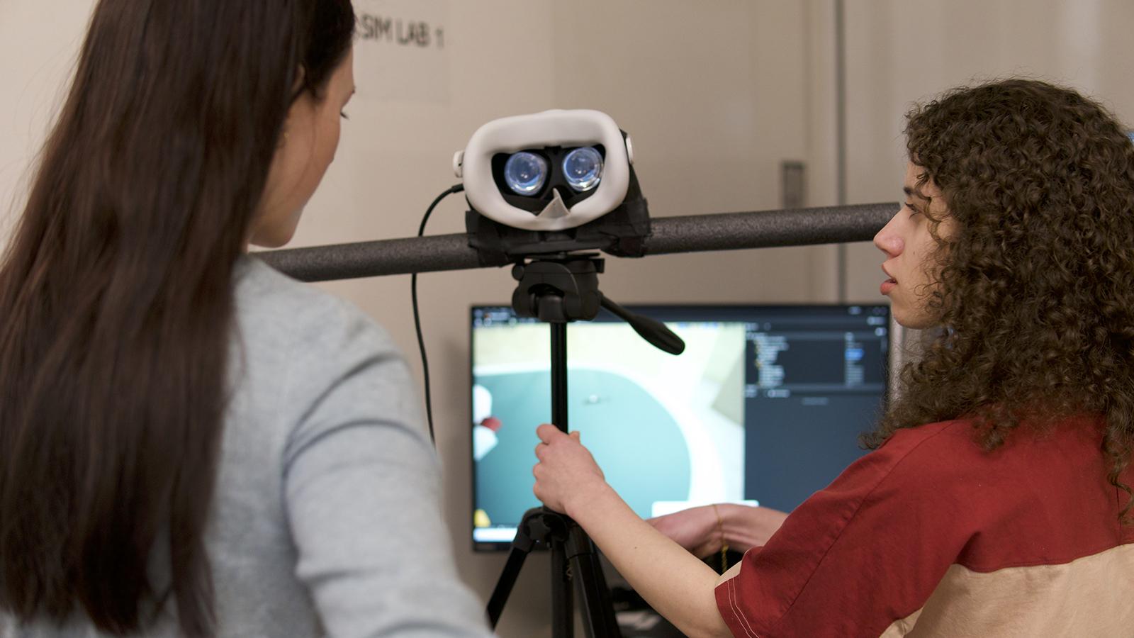 Two people looking at a VR headset that is mounted on a tripod.