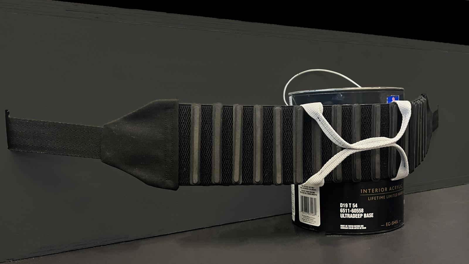 A close-up view of the FrunkFlex cargo-securing system stretched around a paint can. The black elastic strap features ribbed knit padding and a light gray activation handle, demonstrating how it conforms to cargo. The background is a black surface, emphasizing the product’s structure and functionality.