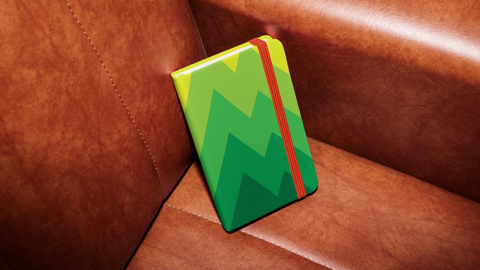 A picture of the climbing journal leaning against a couch seat. The journal itself is various shades of green with a mountain-like pattern. There is an orange elastic strap used to hold the journal closed.