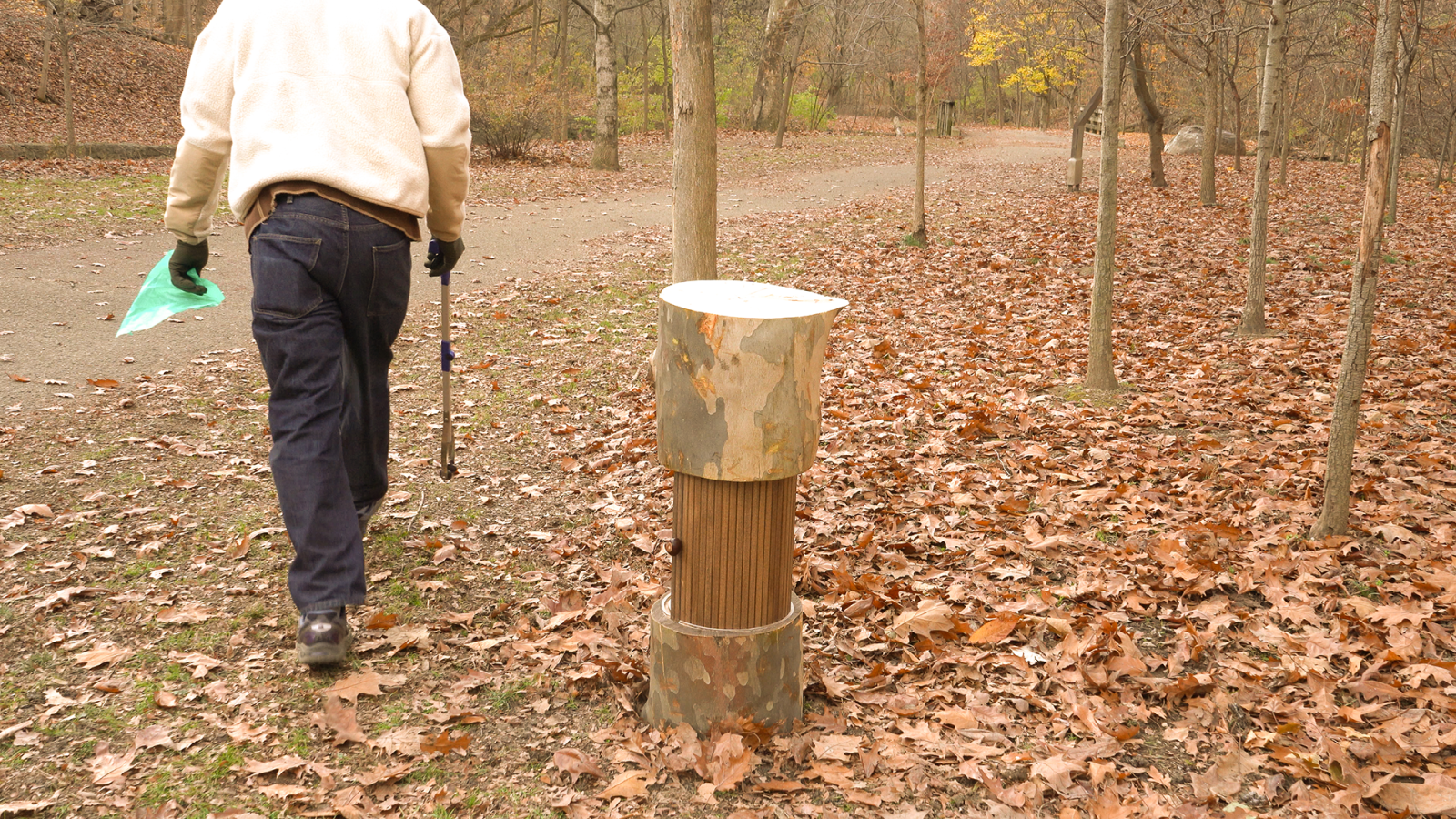 A person walks away from the tree-stump-inspired structure (Heritage Ring), headed toward a paved path with a trash grabber and trash bags.