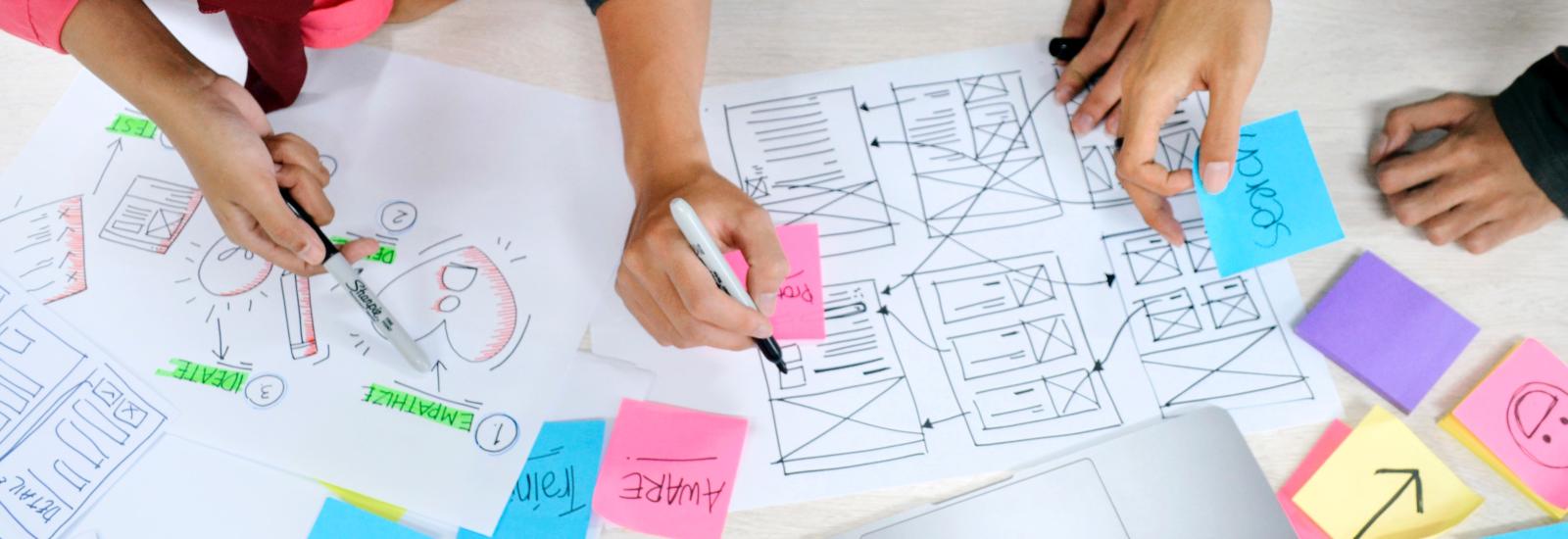 overhead view of three people collaborating at a desk with drawings, post-it notes, and a laptop